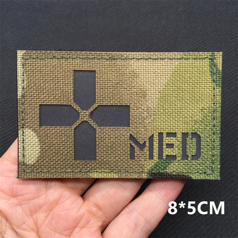 Camouflage medical patch with cross symbol and 'MED' text held in a hand against a dark background.