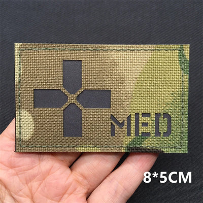 Camouflage medical patch with cross symbol and 'MED' text held in a hand against a dark background.