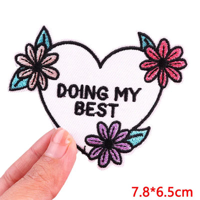 Heart-shaped patch with flowers and 'DOING MY BEST' text held by a hand, on a white background.