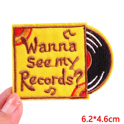 Embroidered patch with 'Wanna See my Records?' text on a yellow background, held by a hand.
