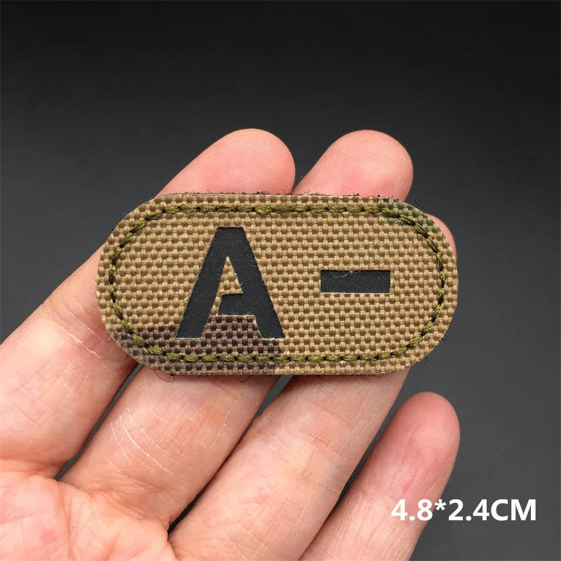 Hand holding an A- multicam IR reflective tactical blood type hook and loop patch approximately 4.8x2.4cm on a dark background