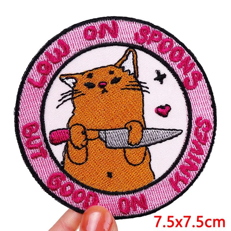 Patch with a cat holding a knife and text on a pink border, held by a hand.