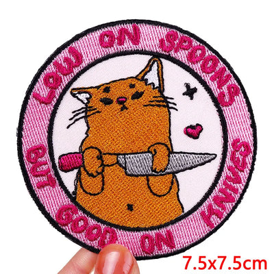 Patch with a cat holding a knife and text on a pink border, held by a hand.