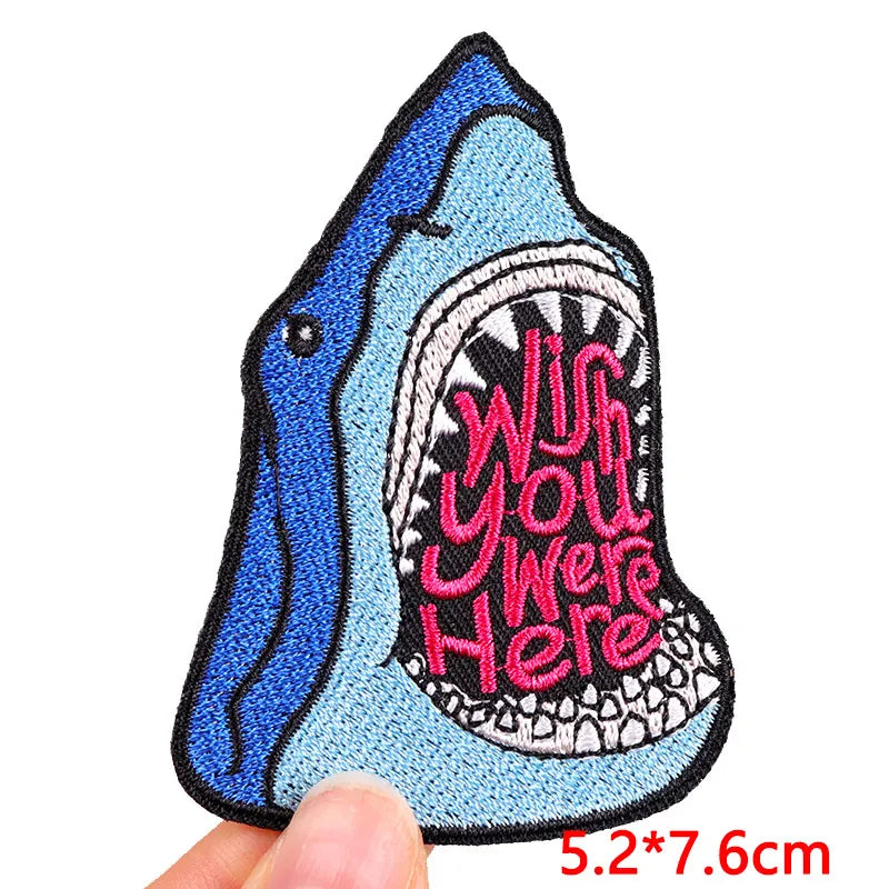 Shark-shaped patch with 'Wish you were here' text held by a hand on a white background