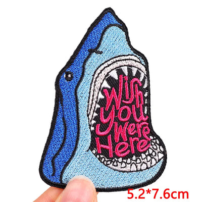 Shark-shaped patch with 'Wish you were here' text held by a hand on a white background