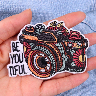 Colorful embroidered patch with a camera design and 'BE YOU TIFUL' text held in a hand.