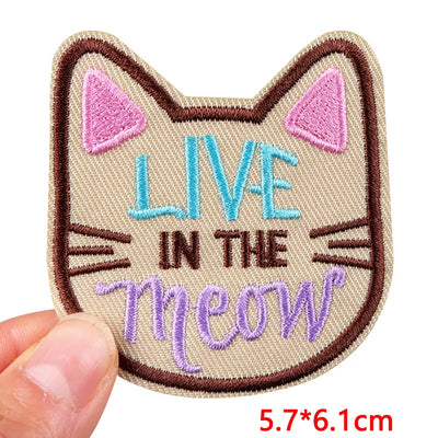 Cat-shaped patch with 'Live in the Meow' text held by a hand, on a white background.