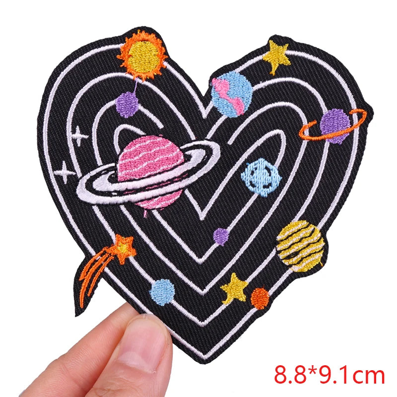 Heart-shaped patch with space-themed design held by a hand, on a white background.