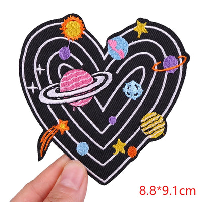 Heart-shaped patch with space-themed design held by a hand, on a white background.
