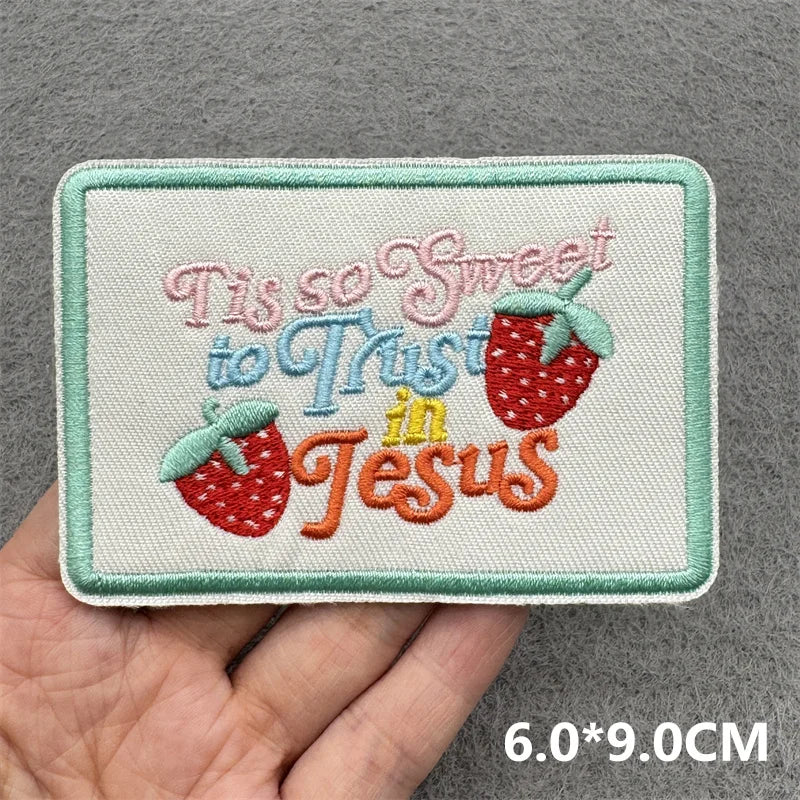 Embroidered patch with 'Tis so sweet to Trust in Jesus' text held in a hand against a gray background.
