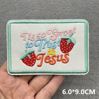Embroidered patch with 'Tis so sweet to Trust in Jesus' text held in a hand against a gray background.