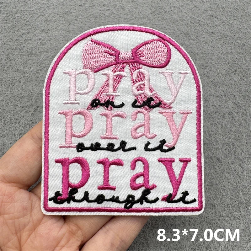 Embroidered patch with 'pray on it pray over it pray though it' text and a pink bow held by a hand on a gray background.