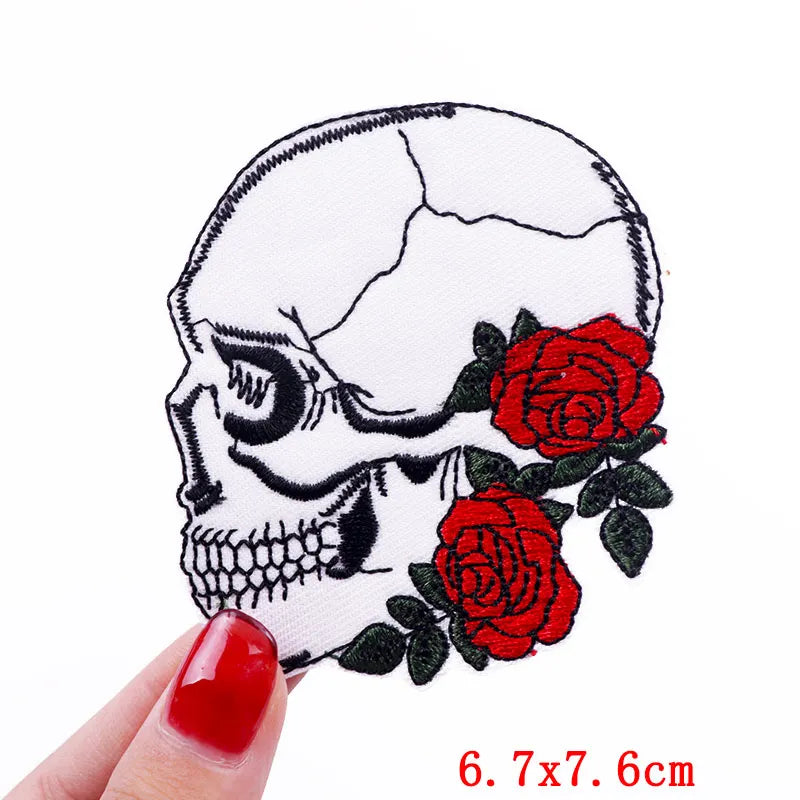 Hand holding an embroidered side-profile skull patch with red roses and dark green leaves approximately 6.7x7.6cm on a white background