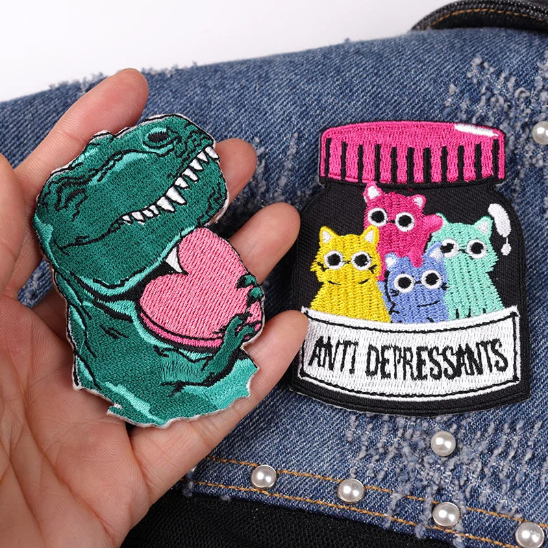 Two colorful patches, one shaped like a dinosaur and the other resembling a bottle labeled 'Anti-Depressants', held over a denim background.