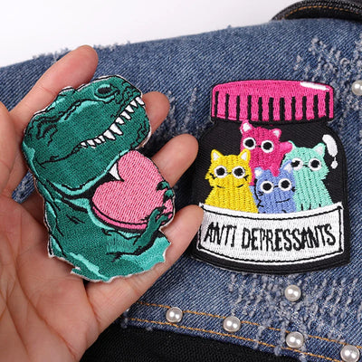 Two colorful patches, one shaped like a dinosaur and the other resembling a bottle labeled 'Anti-Depressants', held over a denim background.