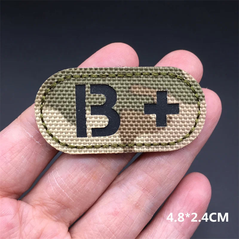 Hand holding an B+ multicam IR reflective tactical blood type hook and loop patch approximately 4.8x2.4cm on a dark background