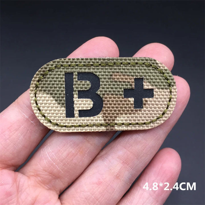 Hand holding an B+ multicam IR reflective tactical blood type hook and loop patch approximately 4.8x2.4cm on a dark background