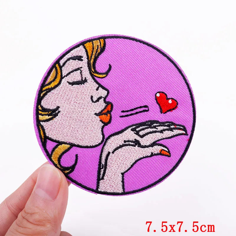 Circular patch with a stylized illustration of a face with blonde hair and a red heart, held by a hand, on a purple background.