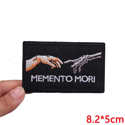 Embroidered patch with 'Memento Mori' text and hands design held by a hand on a white background.