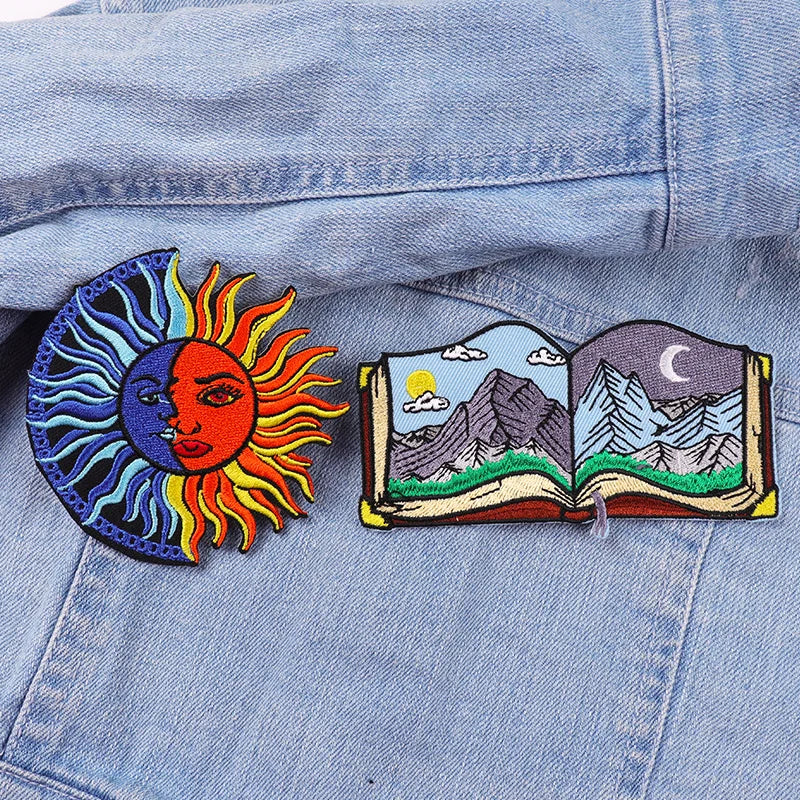 Two embroidered patches on a denim jacket: one sun-like design and one open book with mountain and celestial designs.
