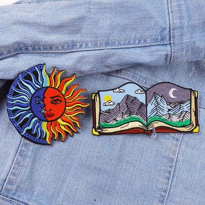 Two embroidered patches on a denim jacket: one sun-like design and one open book with mountain and celestial designs.