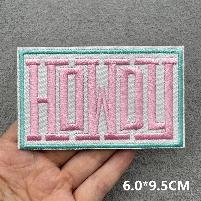 Hand holding a pink and turquoise embroidered Howdy iron-on patch approximately 6x9.5cm on a gray background
