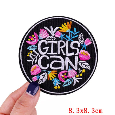 Circular patch with 'GIRLS CAN' text and colorful flowers held by a hand on a white background.