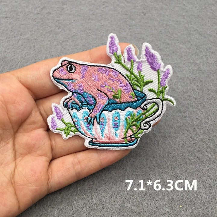Embroidered patch of a frog in a teacup with lavender on a hand, against a gray background.
