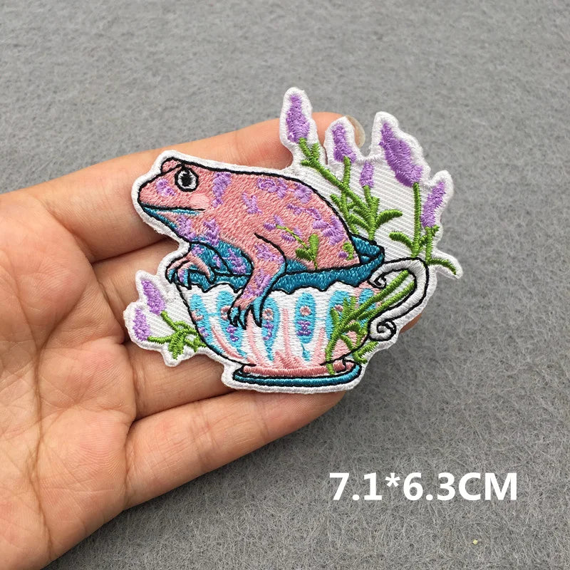Embroidered patch of a frog in a teacup with lavender on a hand, against a gray background.