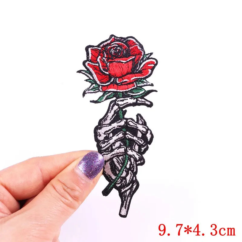 Skeleton hand holding a red rose patch held by a hand with purple nail polish on a white background.