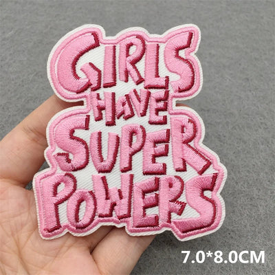 Pink embroidered patch with 'Girls Have Super Powers' text held by a hand on a gray background