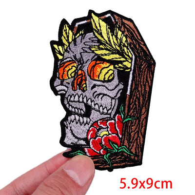 Embroidered patch of a skull in a coffin with roses held by a hand, on a white background.