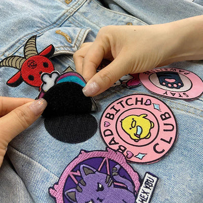 Hand holding a patch over a denim jacket with various patches including a pink 'BITCHO CLUB' patch.