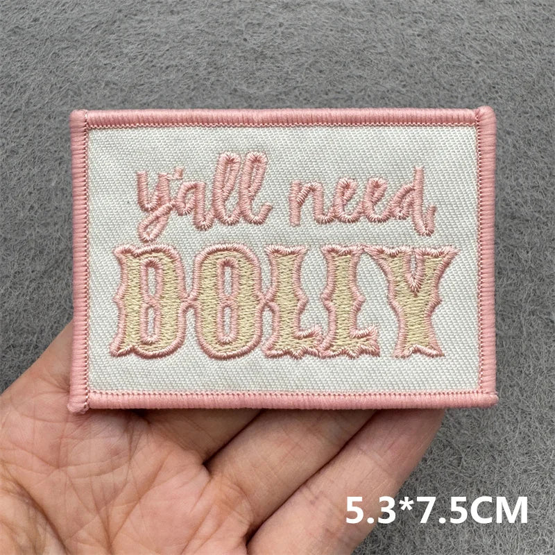 Embroidered patch with 'y'all need DOLLY' text held in a hand against a gray background