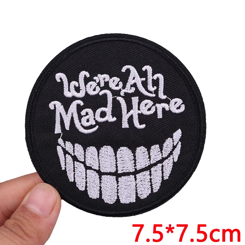Circular patch with 'We're All Mad Here' text and smiley face design held by a hand on a white background.