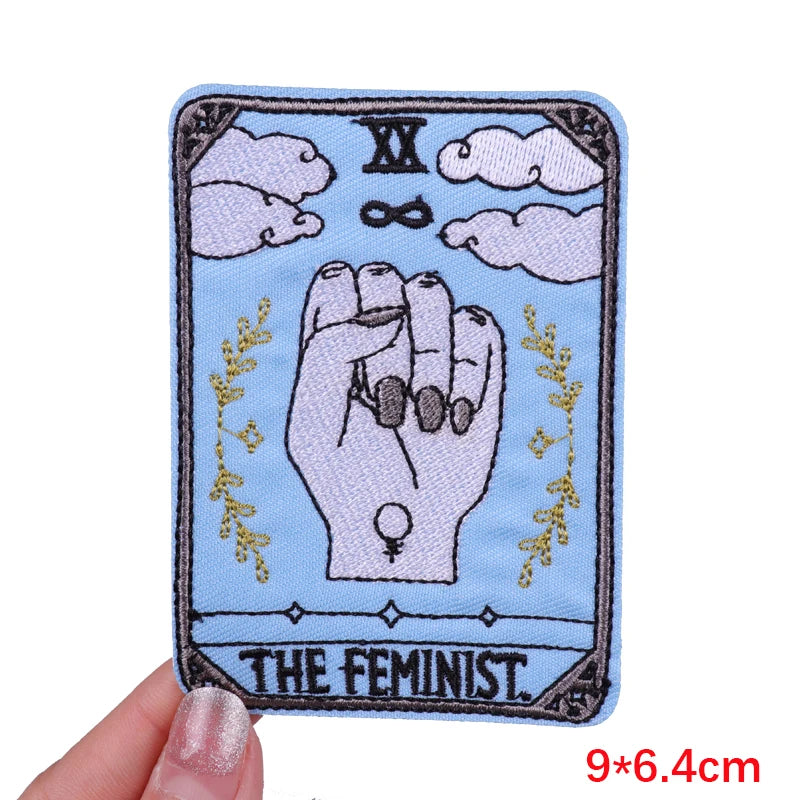 Patch with a hand sign and 'The Feminist' text on a blue background, held by a hand.