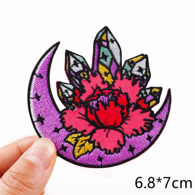 Colorful embroidered patch with a pink flower and purple crescent moon held by a hand on a white background.