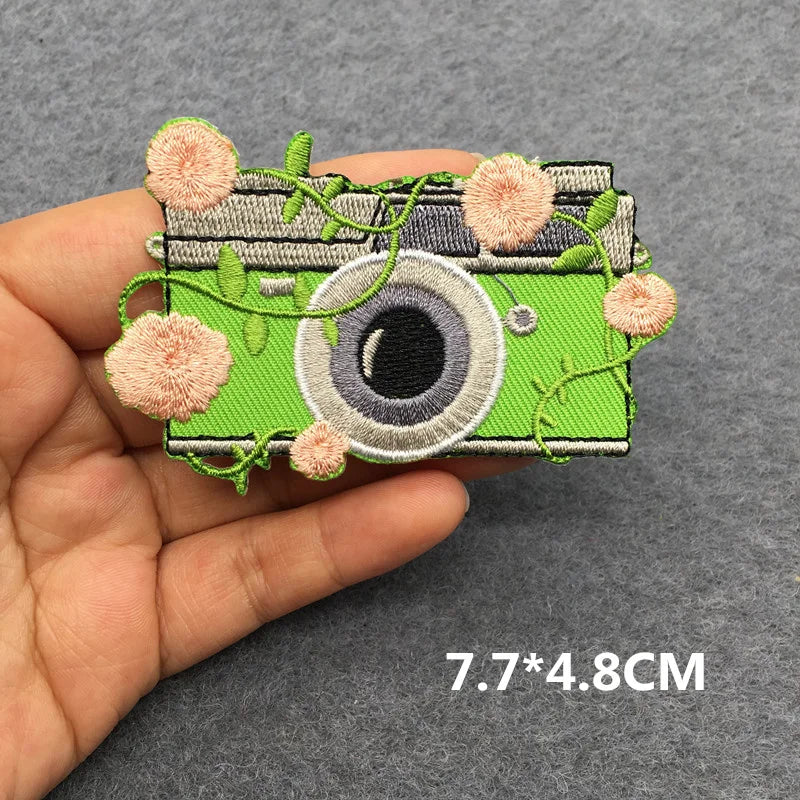 Green camera-shaped patch with floral details held by a hand on a gray background