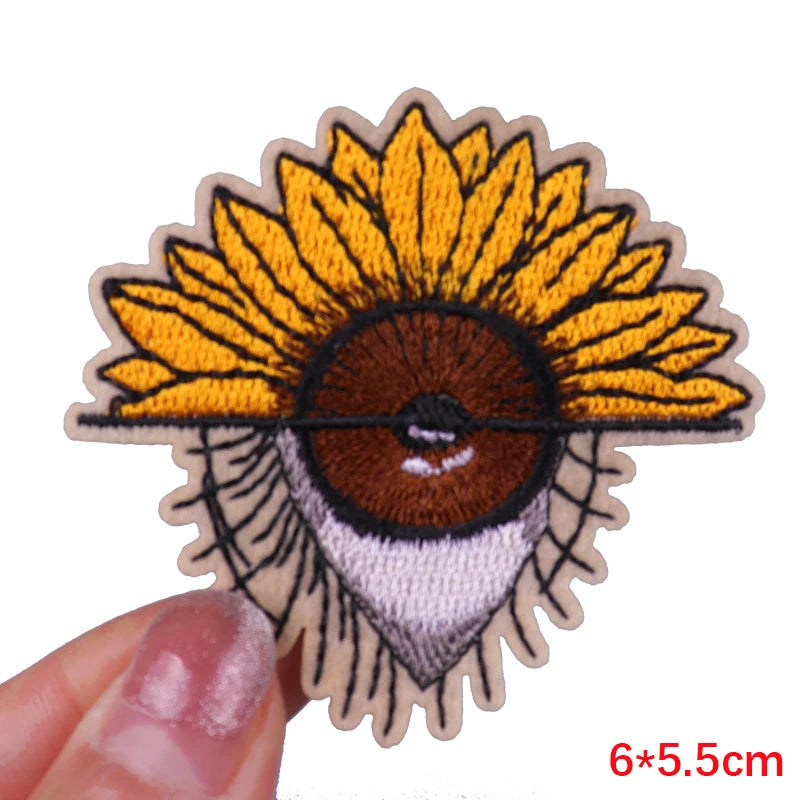 Embroidered patch of a face with a sunflower head held by a hand on a white background.