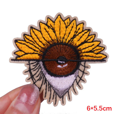 Embroidered patch of a face with a sunflower head held by a hand on a white background.