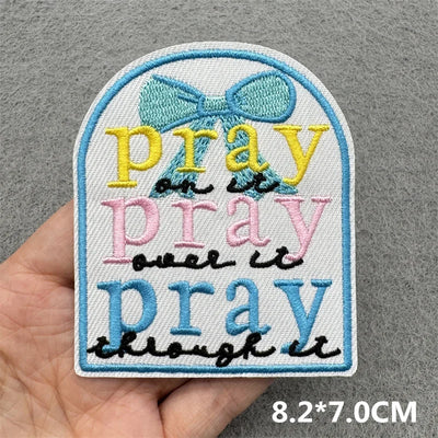 Embroidered patch with 'pray' text and a bow design held by a hand on a gray background.