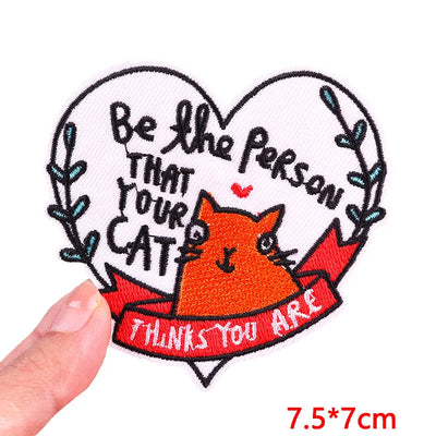 Heart-shaped patch with a cat illustration and text held by a hand, on a white background.