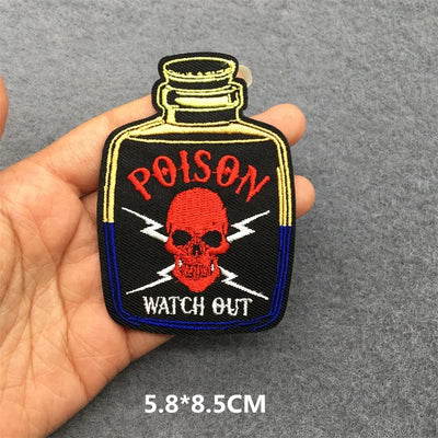 Embroidered patch of a poison bottle with skull design held by a hand on a gray background.