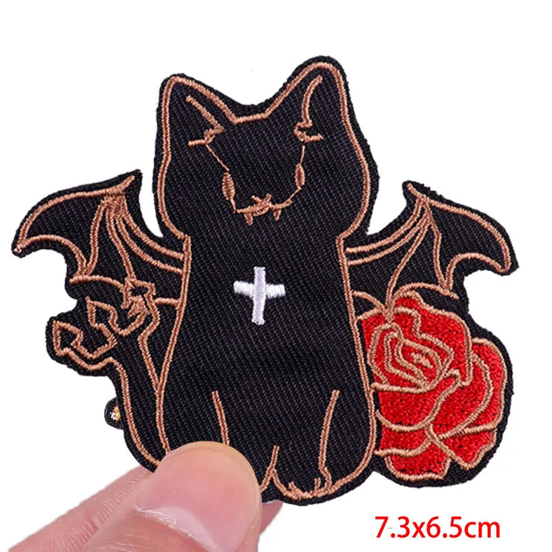 Black cat patch with bat wings and a red rose held by a hand on a white background
