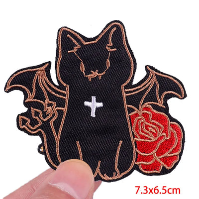 Black cat patch with bat wings and a red rose held by a hand on a white background