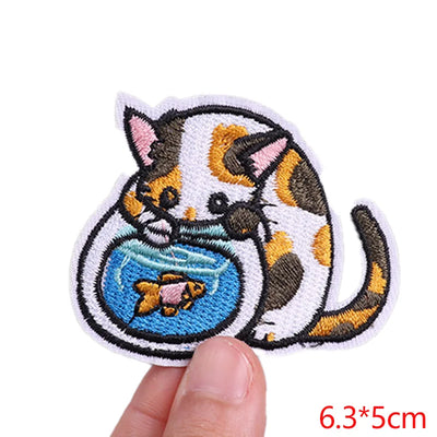 Embroidered patch of a cat with a fishbowl held by a finger, on a white background.