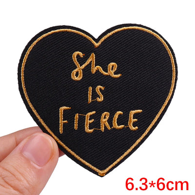 Heart-shaped patch with 'She is Fierce' text held by a hand, on a white background.