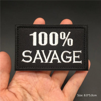 Black patch with '100% SAVAGE' text held in a hand against a dark background