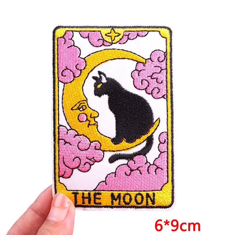 Embroidered patch of 'The Moon' card with a black cat on a yellow crescent moon, held by a hand.