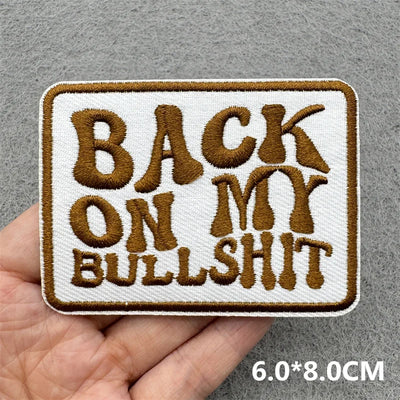 Embroidered patch with text 'BACK ON MY BULLSHIT' held in a hand against a gray background.